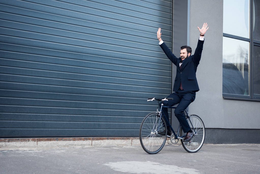 excited young businessman with outstretched arms riding bicycle on street - Photo, Image