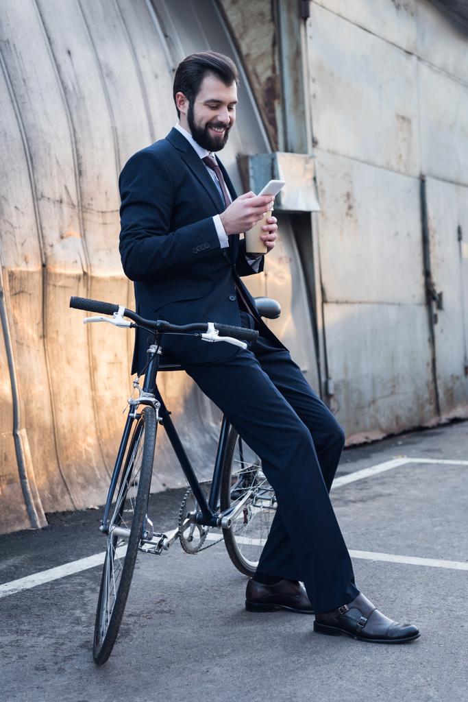 smiling businessman with coffee to go using smartphone while leaning on bicycle on street - Photo, Image