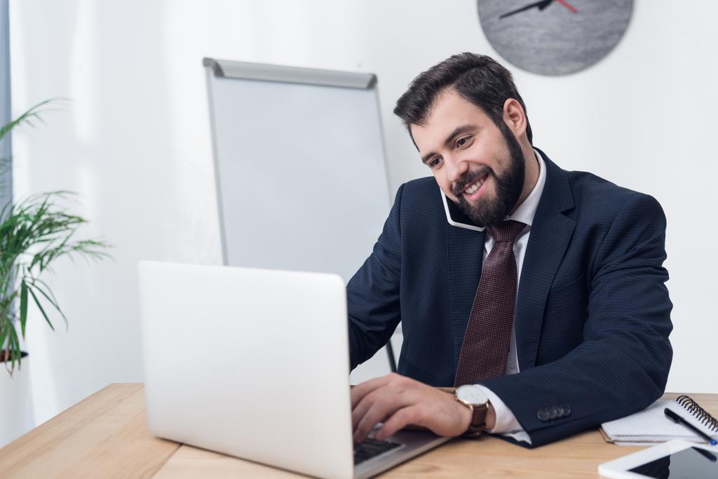 Porträt eines lächelnden Geschäftsmannes, der während der Arbeit am Laptop im Büro mit dem Smartphone spricht - Foto, Bild
