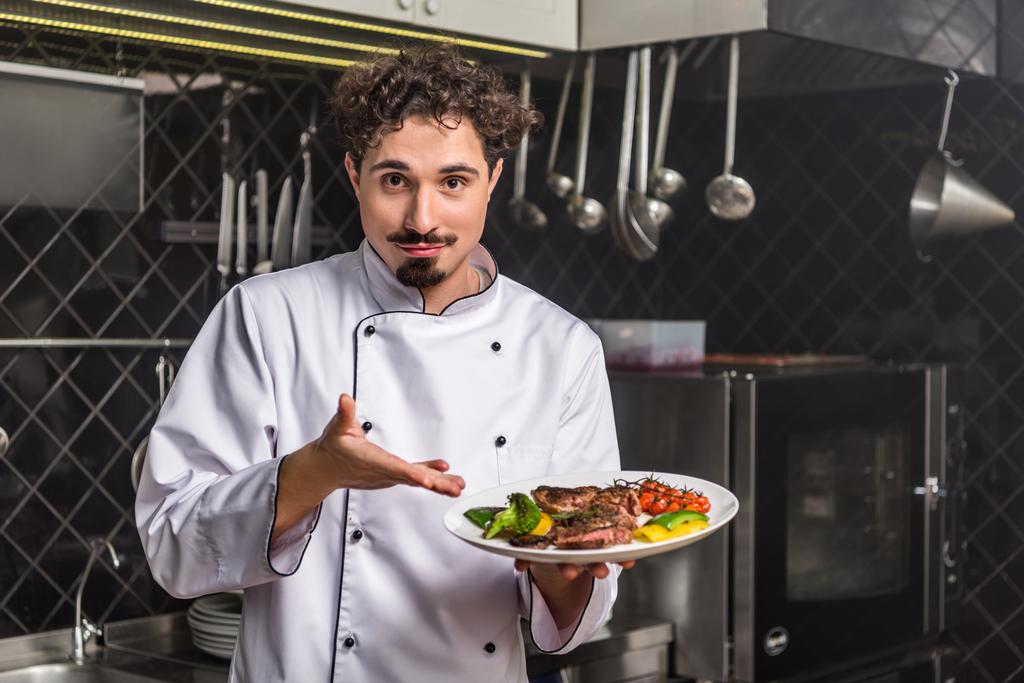 chef bonito mostrando legumes cozidos com carne no prato
 - Foto, Imagem