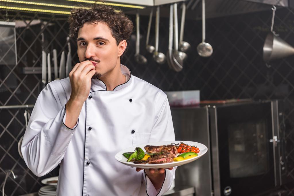 chef segurando prato com legumes fritos e carne e mostrando delicioso sinal
 - Foto, Imagem