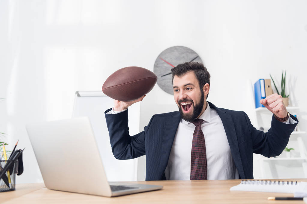 portrait of excited businessman with rugby ball at workplace with laptop in office - Photo, Image
