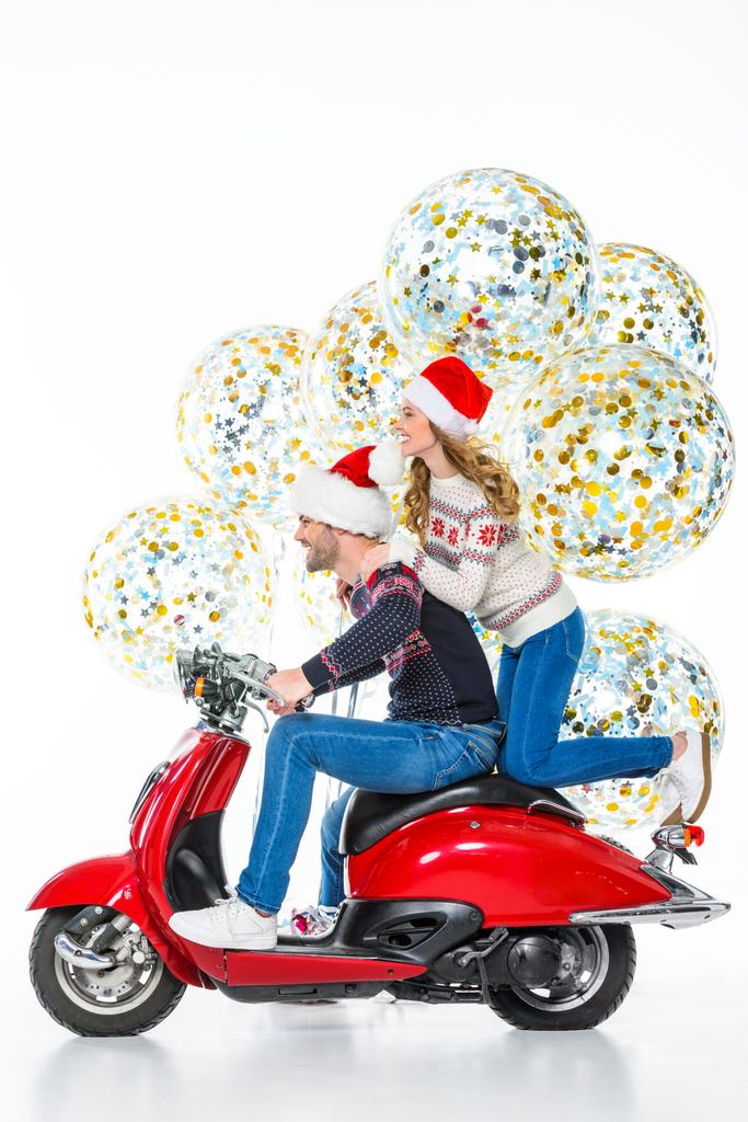 cheerful couple in santa hats riding red scooter, big christmas balloons with confetti on background, isolated on white  - Photo, Image