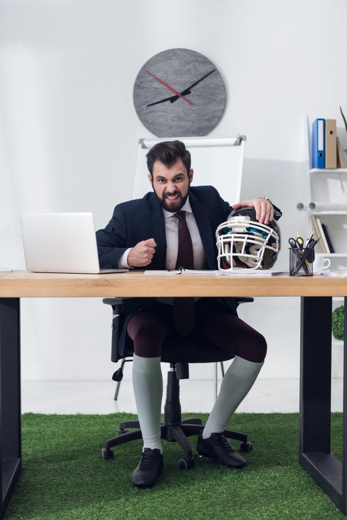 empresario enojado sentado en el lugar de trabajo con casco de rugby en la oficina
 - Foto, imagen