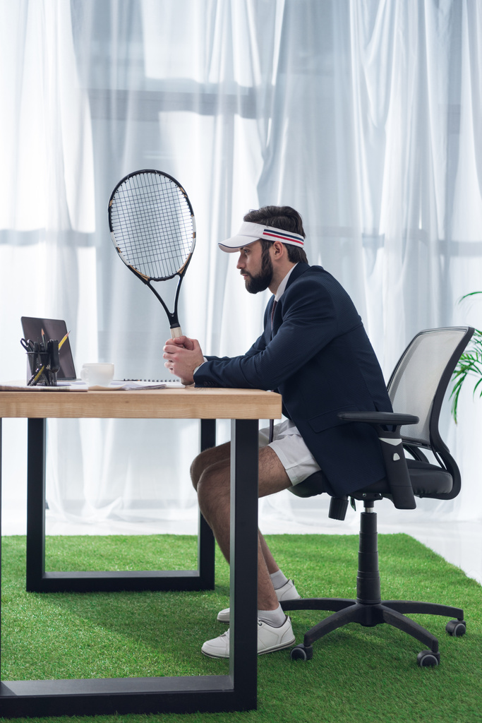 боковой вид бизнесмена в кепке с теннисной ракеткой на рабочем месте в офисе
 - Фото, изображение