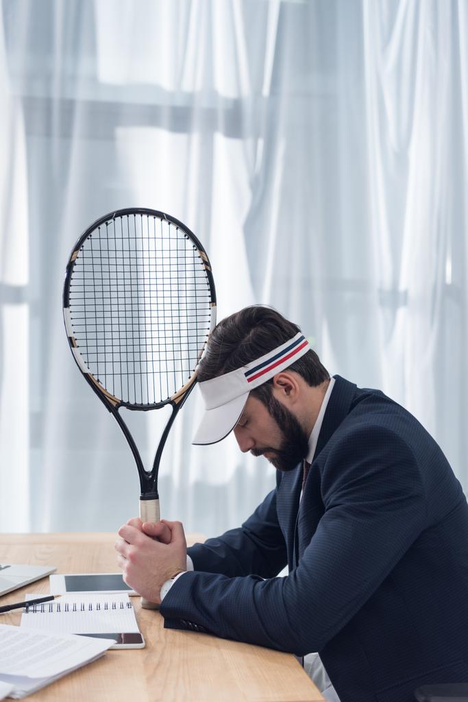 Zijaanzicht van peinzende zakenman in pak met een tennisracket op werkplek - Foto, afbeelding