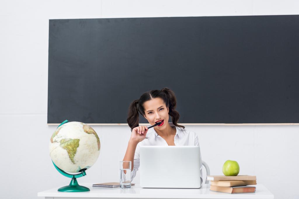 young seductive teacher sitting at workplace with globe and laptop in classroom - Photo, Image