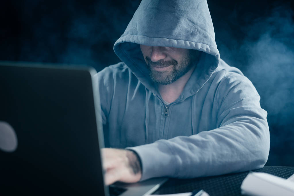 secretive mysterious man hacker hooded hides his face doing something illegal on a laptop smiling - Photo, Image
