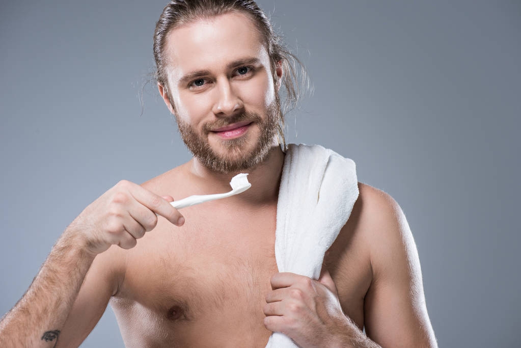 Smiling bearded man with towel on shoulder holding toothbrush with toothpaste in hand, isolated on gray   - Photo, Image