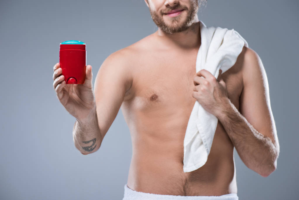 Sonriente hombre medio desnudo sosteniendo toalla de baño sobre el hombro en una mano y palo desodorante en otra, aislado en gris
 - Foto, Imagen