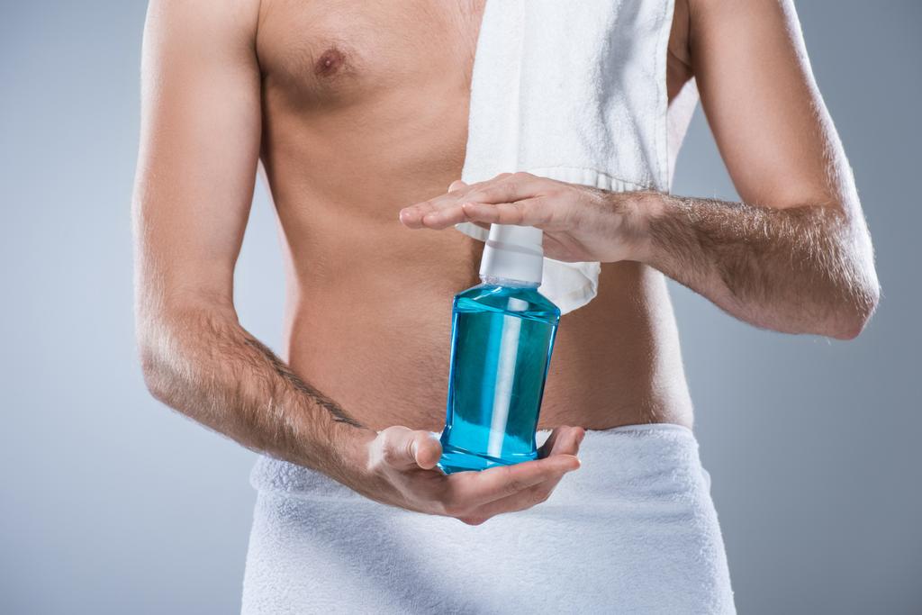 Midsection of man with bath towel on shoulder and around waist holding bottle of tooth rinse in hands, isolated on gray  - Photo, Image