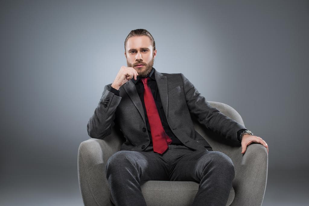 serious young businessman in formal suit sitting on armchair, with hand on chin, isolated on gray - Photo, Image