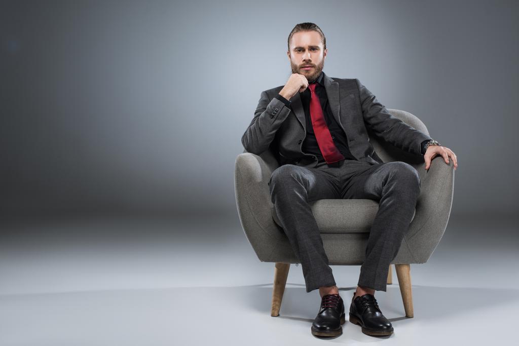 Joven empresario serio sentado en sillón con la mano a la barbilla y mirando a la cámara, aislado en gris
 - Foto, Imagen