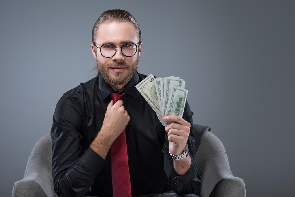 Succesvolle Glimlachende man met geld in de hand zitten in een leunstoel tijdens het strekken van de stropdas, geïsoleerd op grijs - Foto, afbeelding