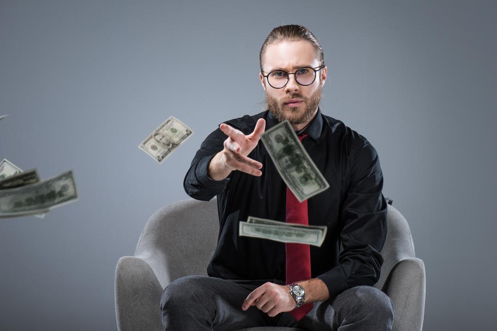 Giovane uomo d'affari di successo in occhiali seduto in poltrona mentre gettava soldi alla macchina fotografica, isolato su grigio
 - Foto, immagini