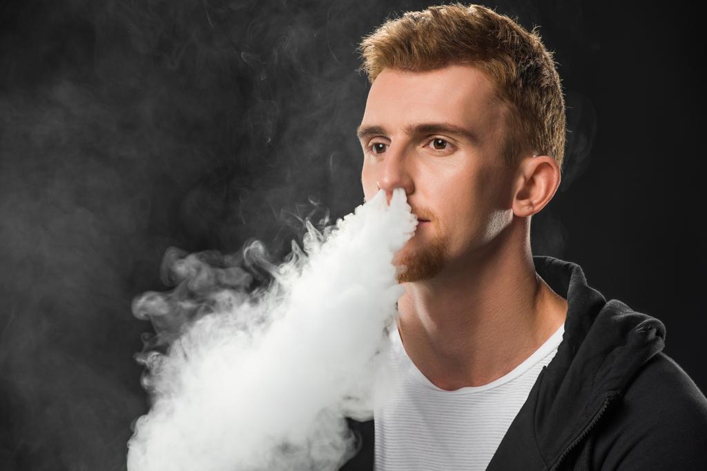 junger bärtiger Mann atmet Rauch elektronischer Zigarette ein, umgeben von Dampfwolken - Foto, Bild