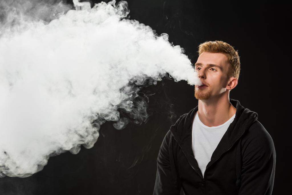 Молодий бородатий чоловік видихає дим електронної сигарети в оточенні хмар пари
 - Фото, зображення