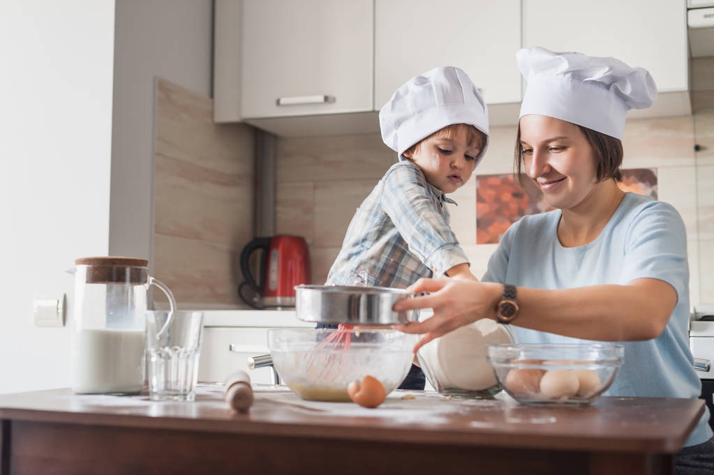 счастливая молодая мать и ребенок в шляпах шеф-повара готовят тесто на кухне
 - Фото, изображение