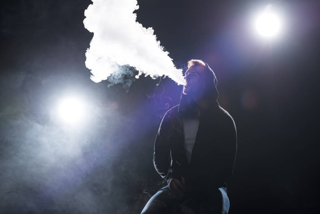 Jeune homme barbu expirant fumée de cigarette électronique contre rétro-éclairé
 - Photo, image