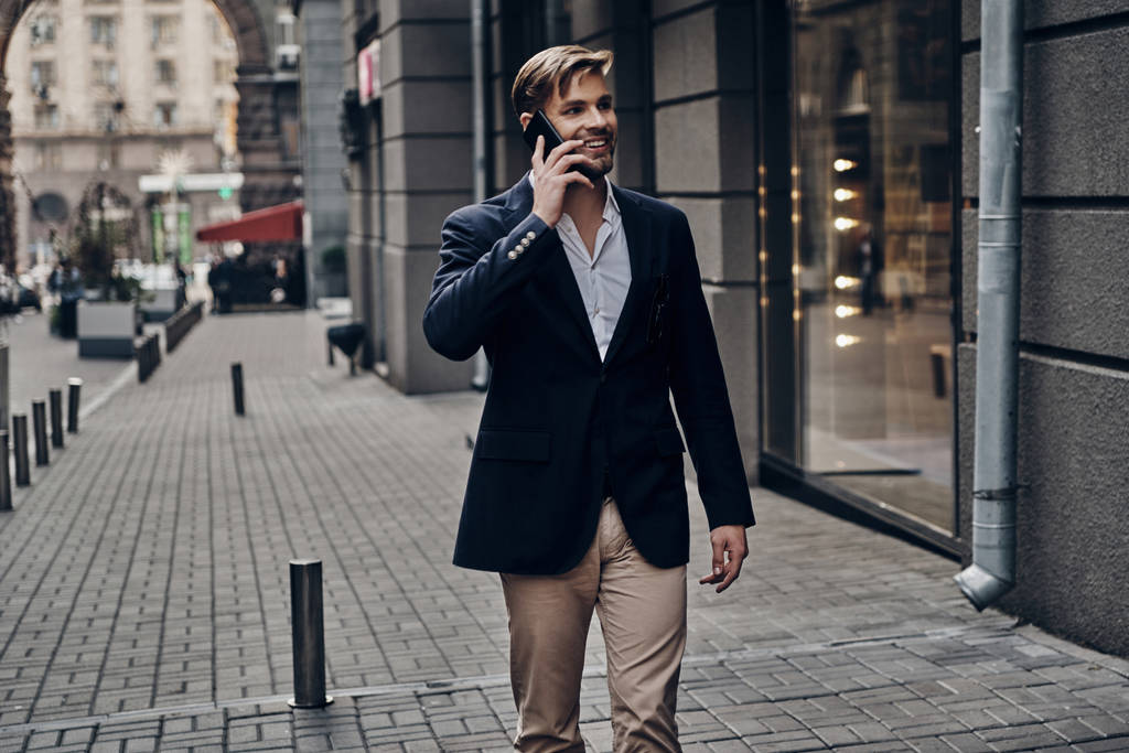knappe jongeman in smart casual dragen op slimme telefoon praten en lopen in de stad straat - Foto, afbeelding