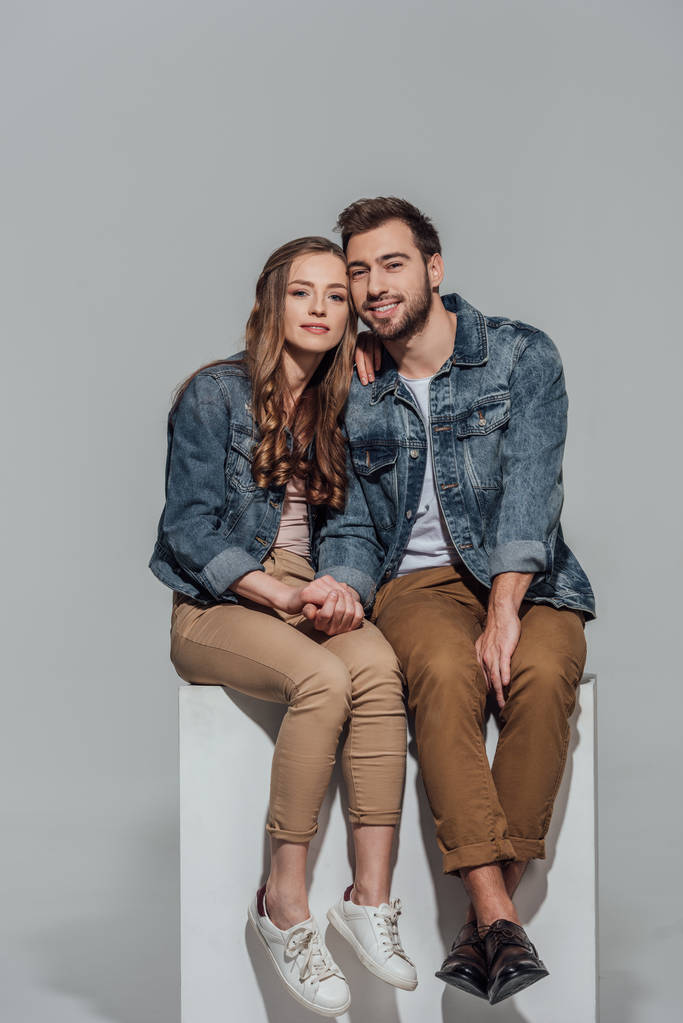 όμορφο νεαρό ζευγάρι στο τζιν μπουφάν κάθονται μαζί και να χαμογελά στη φωτογραφική μηχανή που απομονώνονται σε γκρι - Φωτογραφία, εικόνα