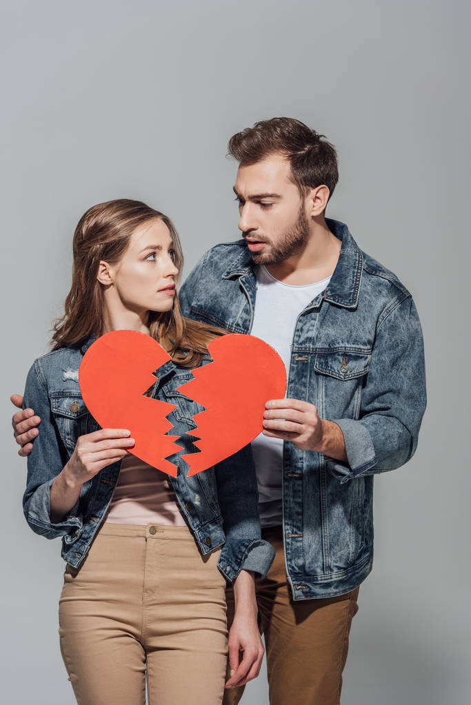 upset young couple holding pieces of broken heart symbol and looking at each other isolated on grey   - Photo, Image