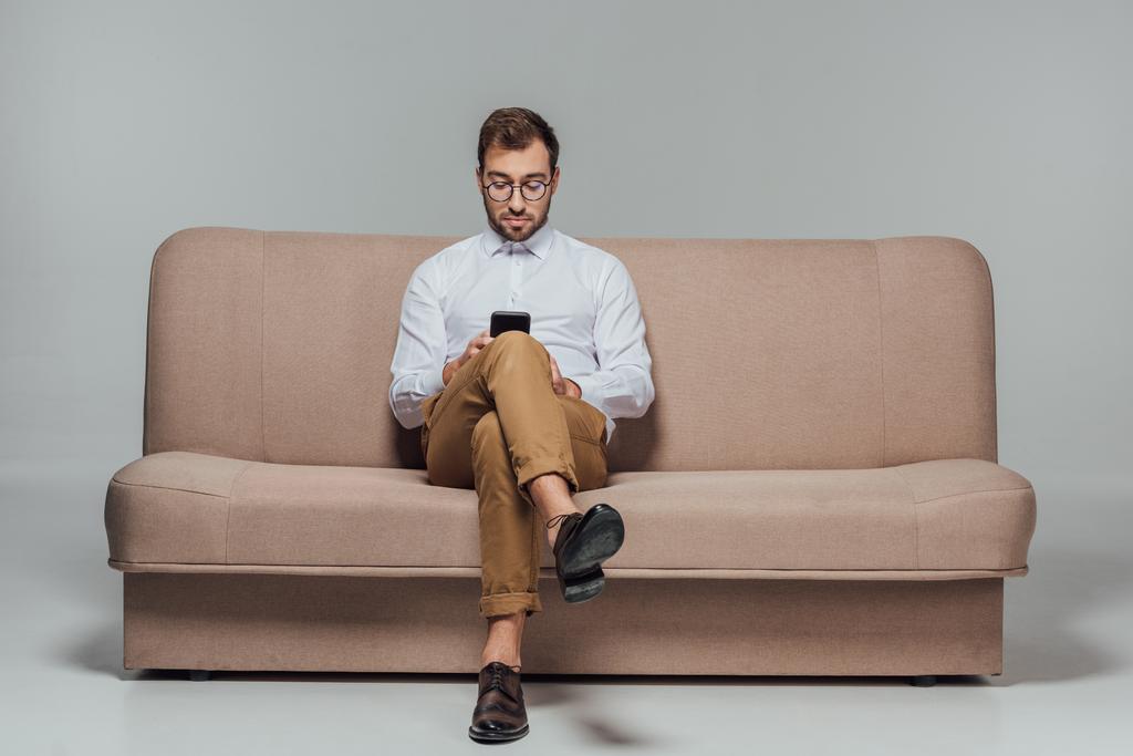 стильный мужчина в очках сидит на диване и использует смартфон, изолированный на сером
 - Фото, изображение