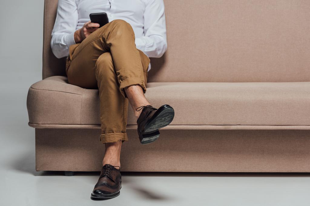 lage sectie van jonge man met behulp van smartphone zittend op de Bank geïsoleerd op grijs - Foto, afbeelding