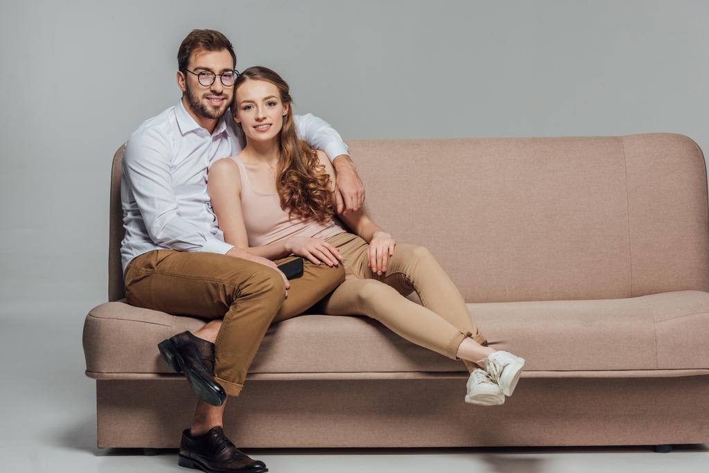 Happy elegantní mladý pár s smartphone se usmívá na kameru při seděli spolu na pohovce izolované Grey - Fotografie, Obrázek
