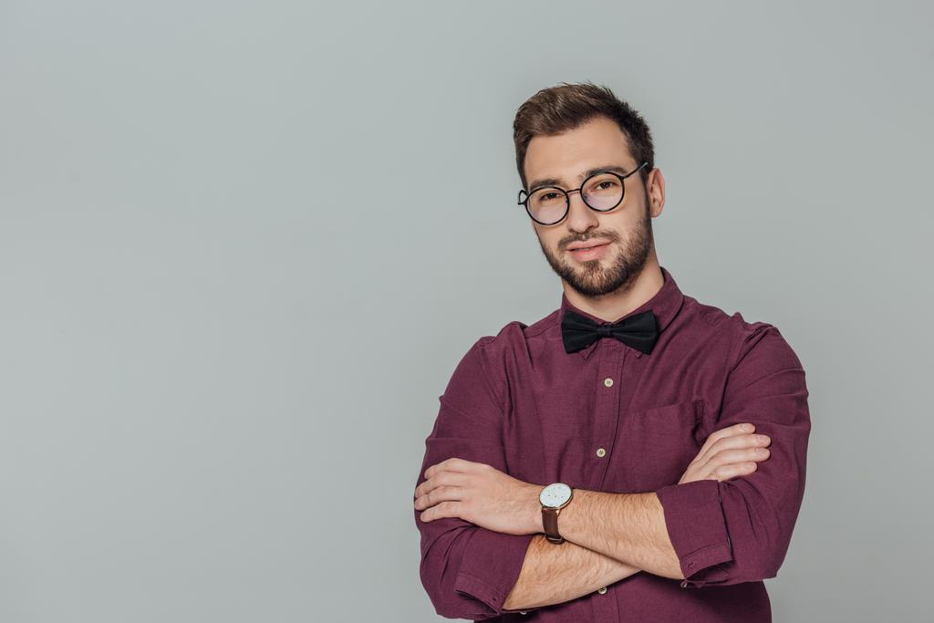 stylish young man in eyeglasses standing with crossed arms and smiling at camera isolated on grey  - Photo, Image