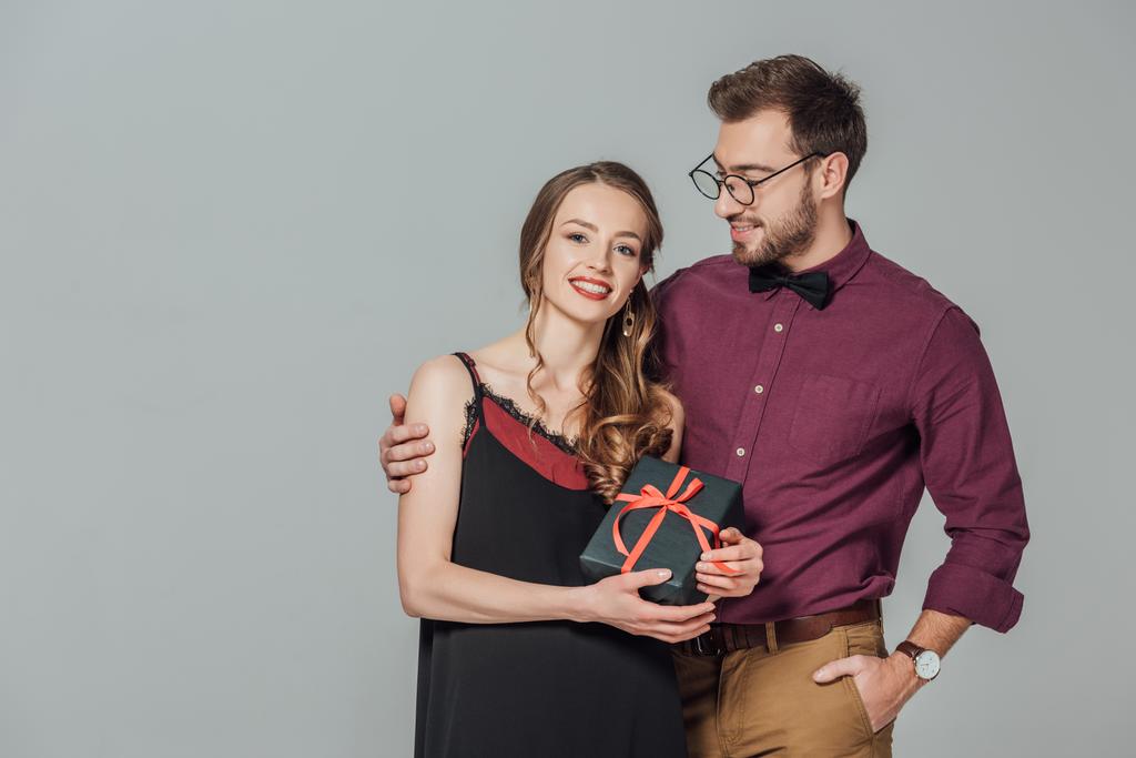 guapo elegante hombre sonriendo y abrazando hermosa mujer joven sosteniendo caja de regalo aislado en gris
 - Foto, imagen