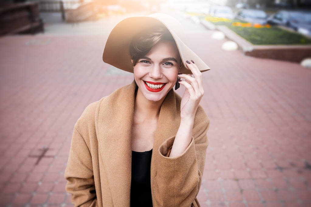 Chica con un abrigo marrón un sombrero marrón está caminando y posando en los interiores de la ciudad. La chica está sonriendo, revisando su teléfono inteligente y tomando café de la cafetería para llevar
. - Foto, Imagen