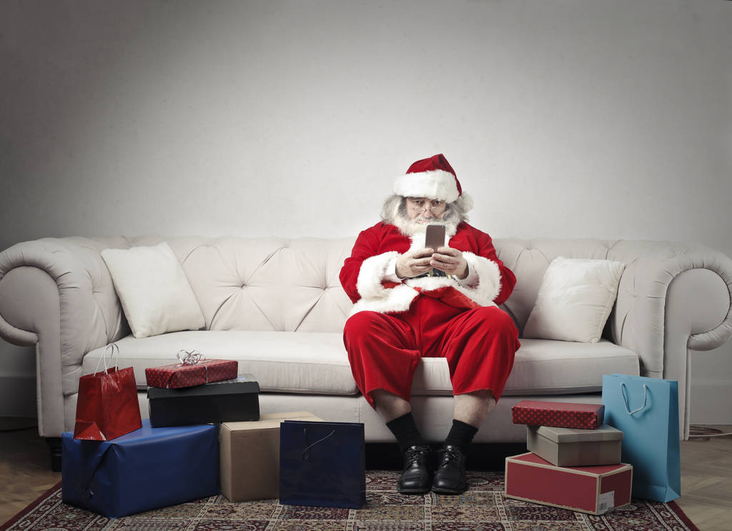 Santa Claus sentado en un sofá con un teléfono inteligente en sus manos y algunos regalos alrededor
 - Foto, Imagen