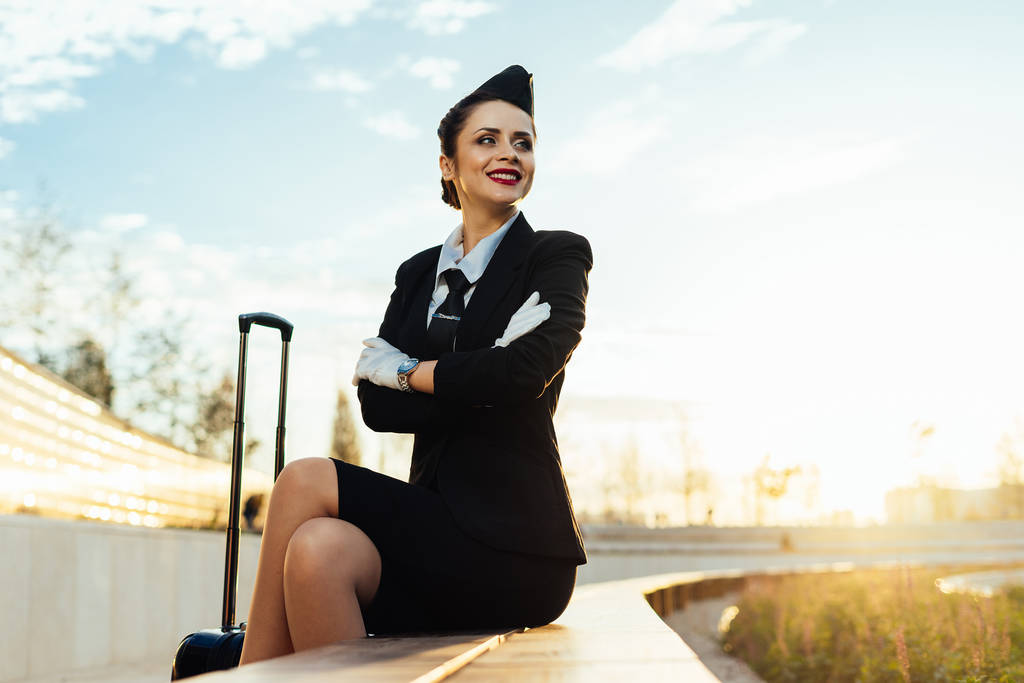 スーツケースの彼女の飛行を待っていると制服を着た幸せの笑みを浮かべて女性スチュワーデス - 写真・画像