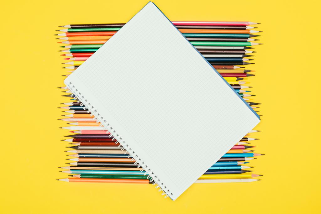Κάτοψη της σύνθεσης των πολύχρωμα μολύβια με κενό σημειωματάριο που απομονώνονται σε κίτρινο φόντο - Φωτογραφία, εικόνα