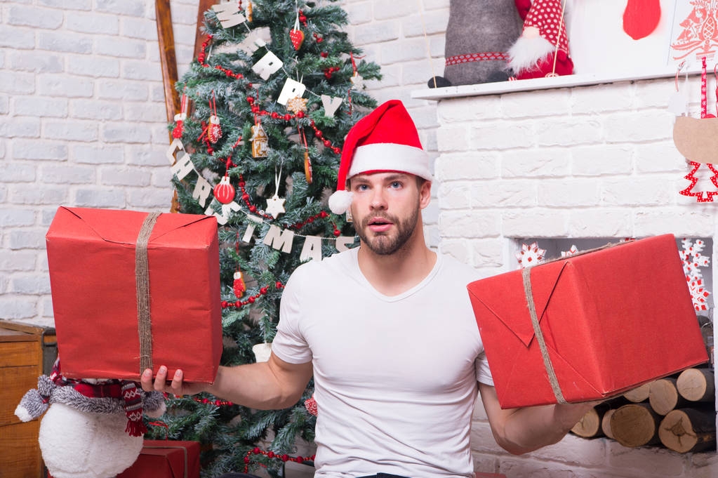 Ο άνθρωπος με τα κόκκινα κουτιά στο χριστουγεννιάτικο δέντρο. Macho στο santa καπέλο κρατήστε τυλιγμένα δώρα στο τζάκι. Καλά Χριστούγεννα και Ευτυχισμένο το νέο έτος. Boxing day έννοια. Διακοπές σε προετοιμασία και γιορτή. - Φωτογραφία, εικόνα