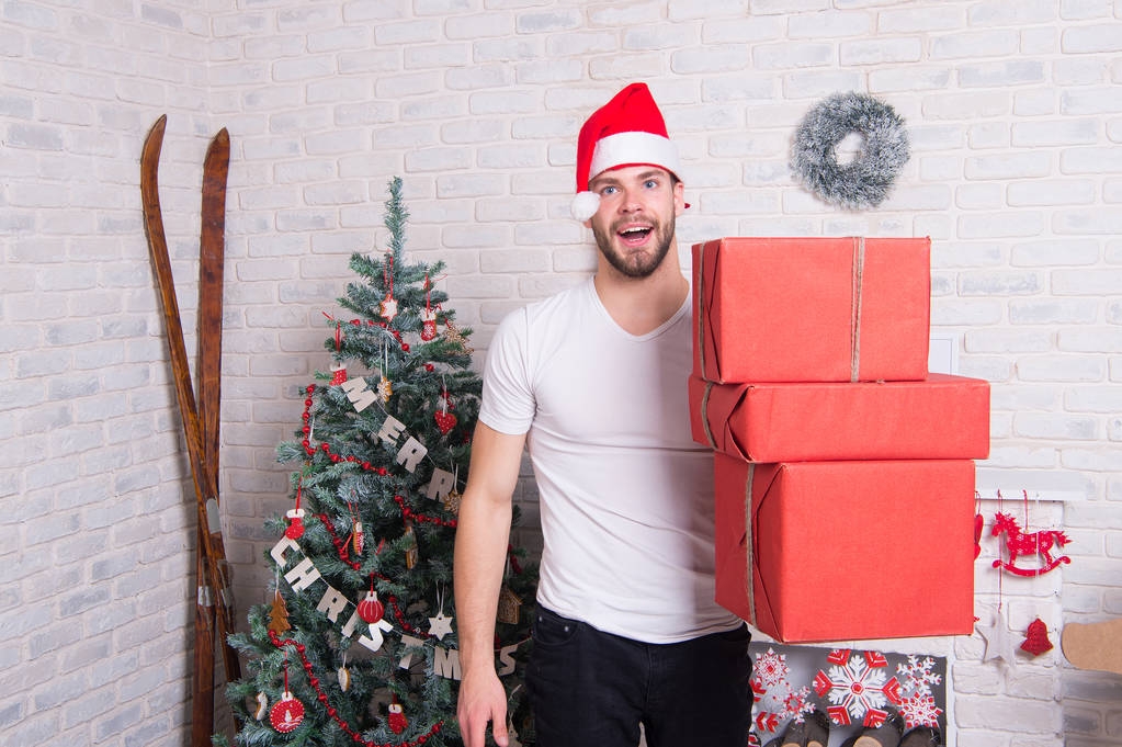 Mann Weihnachtsmann lächelt mit Schachteln am Weihnachtsbaum. Schenken und Tauschen. Feiertagsvorbereitung und -feier. Macho mit rotem Hut hält verpackte Geschenke in der Hand. Boxtag-Konzept. - Foto, Bild