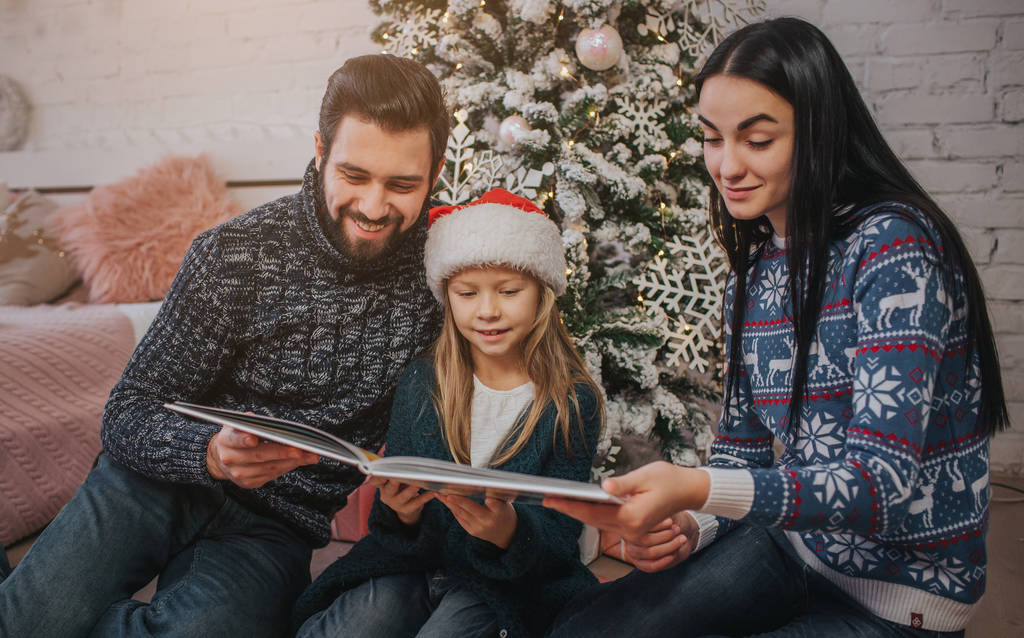 Hyvää joulua ja Happy Holidays Iloinen äiti, isä ja hänen söpö tytär tyttö lukee kirjaa. Vanhemmat ja pieni lapsi hauskaa lähellä joulukuusi sisätiloissa. Huomenta, joulu. Muotokuva perhe lähikuva
 - Valokuva, kuva
