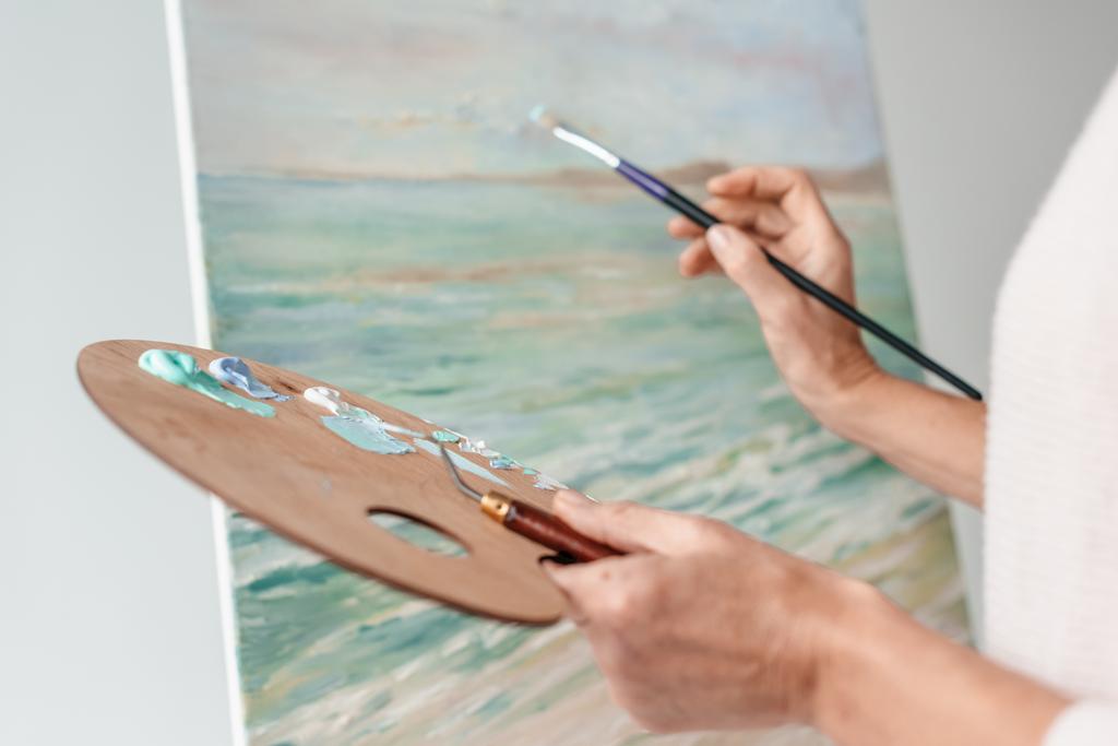 recortado tiro de artista sosteniendo paleta y pincel mientras que la pintura en el caballete en el estudio de arte
  - Foto, Imagen