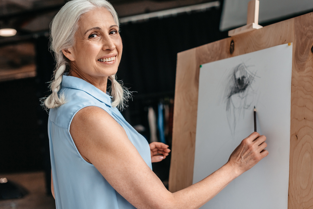 hermosa mujer mayor sonriendo a la cámara mientras dibuja con lápiz en caballete en la clase de arte
 - Foto, Imagen