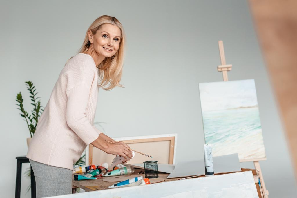 beautiful mature woman smiling at camera while holding art tools in art studio - Photo, Image