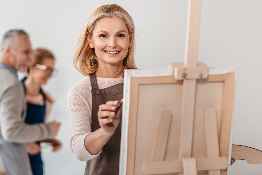 bela mulher madura sorrindo para a câmera enquanto pintura no cavalete na aula de arte
  - Foto, Imagem