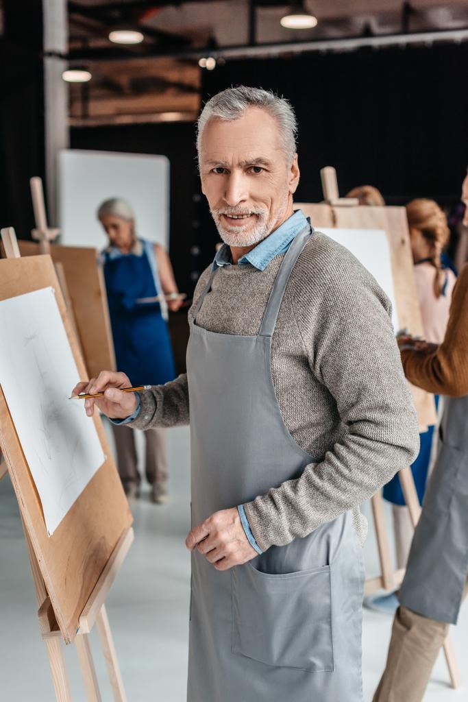 vanhempi mies hymyilee kameralle seisoessaan lähellä maalaustelinettä taidetunnilla
 - Valokuva, kuva