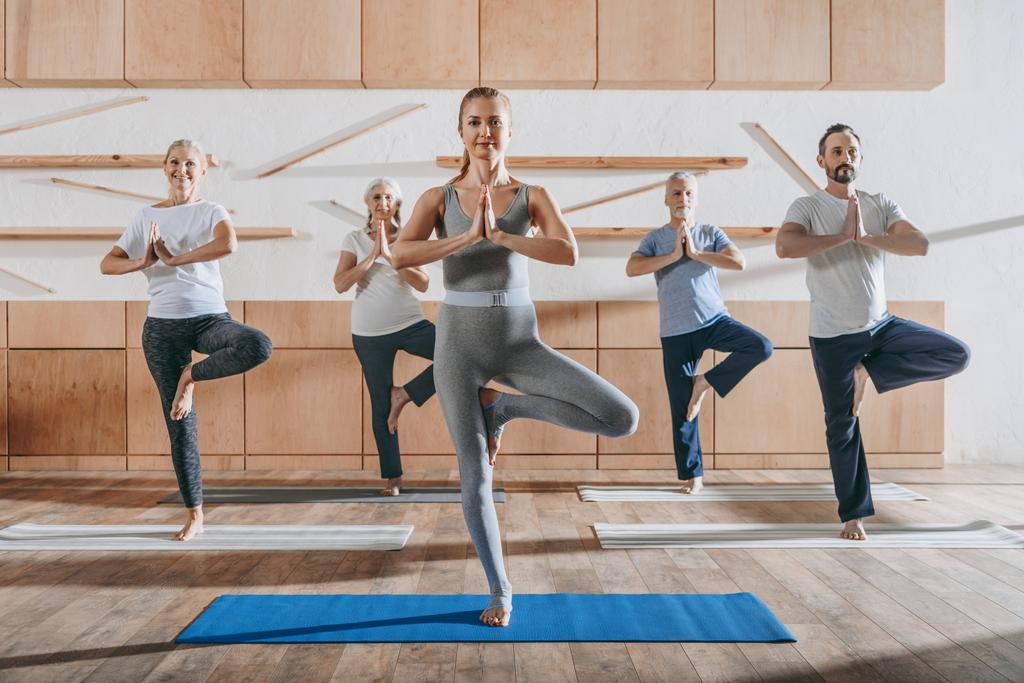 grupo de pessoas idosas praticando ioga com instrutor em pose de árvore em tapetes em estúdio
 - Foto, Imagem