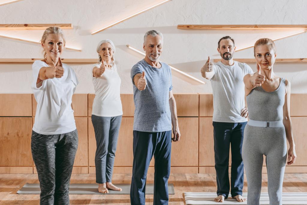 Senior mensen en instructeur in sportkleding duimen opdagen en glimlachend in de camera in de fitness-studio - Foto, afbeelding