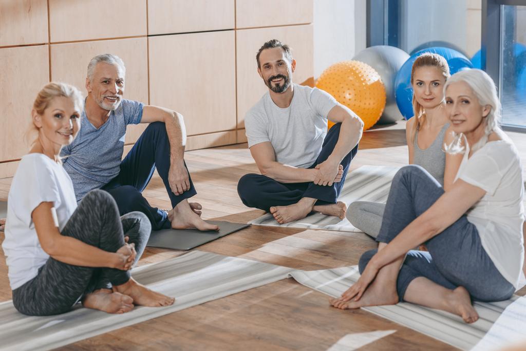 volwassen mensen met instructeur zittend op yoga matten en camera te kijken - Foto, afbeelding