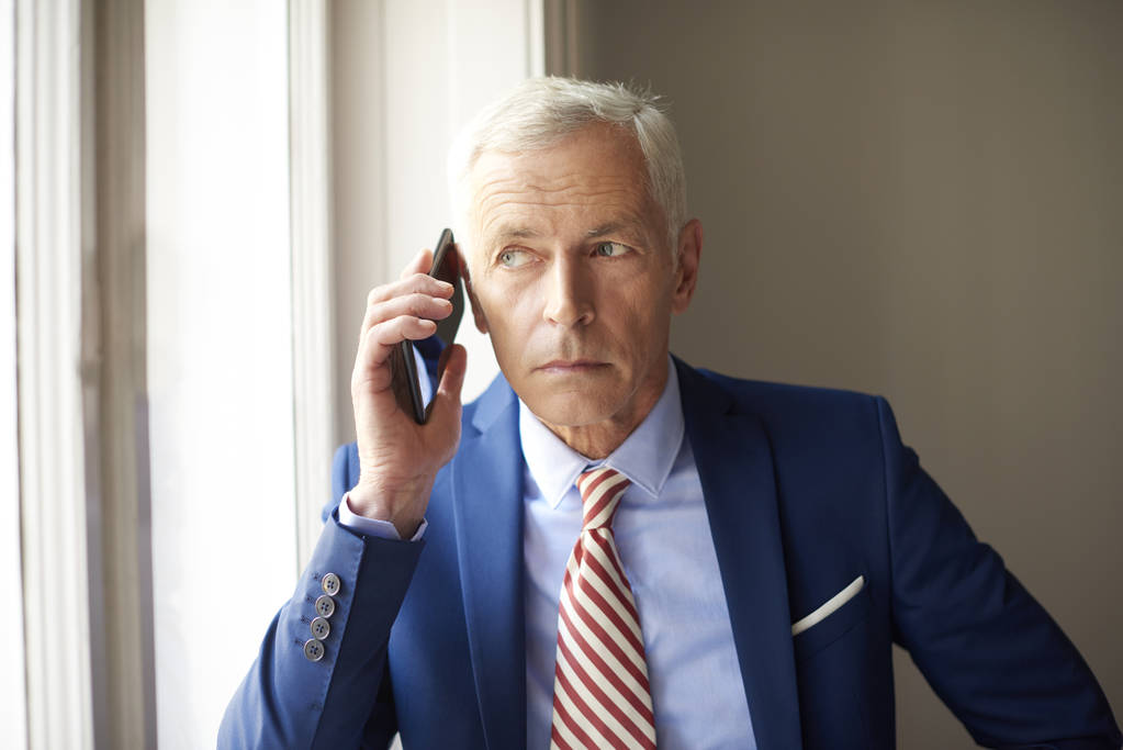 Ανώτερος επιχειρηματίας χρησιμοποιώντας το κινητό του τηλέφωνο και κάνοντας κλήση ενώ στέκεται στο παράθυρο  - Φωτογραφία, εικόνα