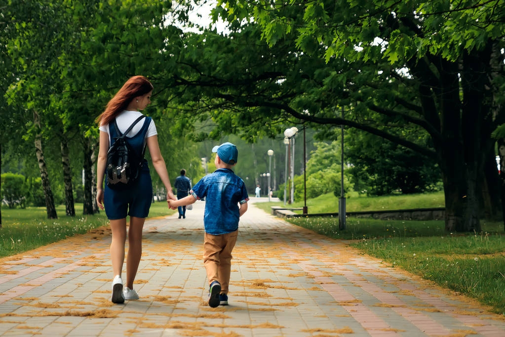 Nuori äiti ja hänen poikansa kävelevät pitkin kaunista kesäpuistoa. Poika ja kaunis nainen kävelevät ja pitävät hauskaa. Perheen muotokuva ulkona
. - Valokuva, kuva