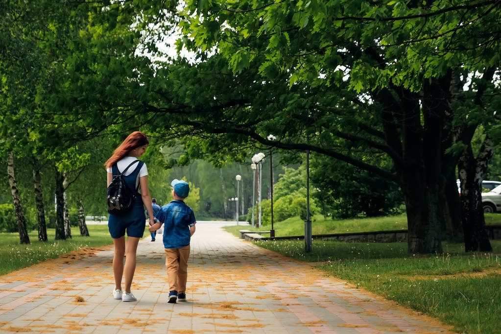 Una giovane madre e suo figlio stanno camminando lungo il bellissimo parco estivo. Un ragazzo e una bella donna camminano e si divertono. Ritratto di famiglia all'aperto
. - Foto, immagini