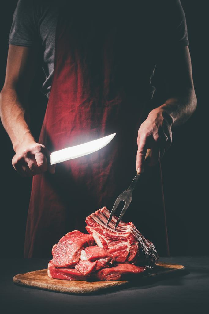 butcher with knife and fork cutting raw meat on wooden cutting board - Photo, Image